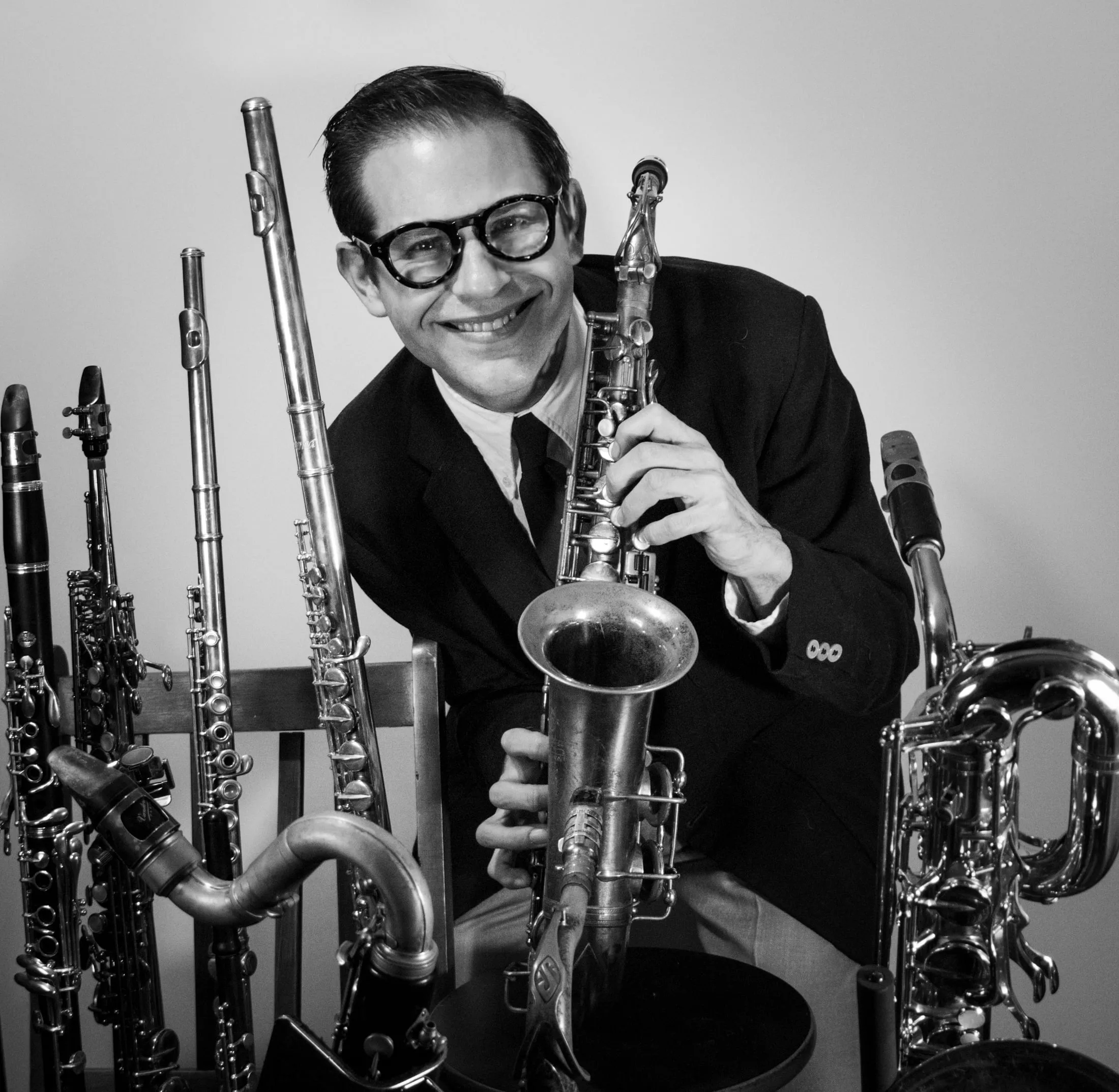 Aaron Johnson grinning as he leans over a saxophone, his full rack of woodwinds behind him, in black and white.